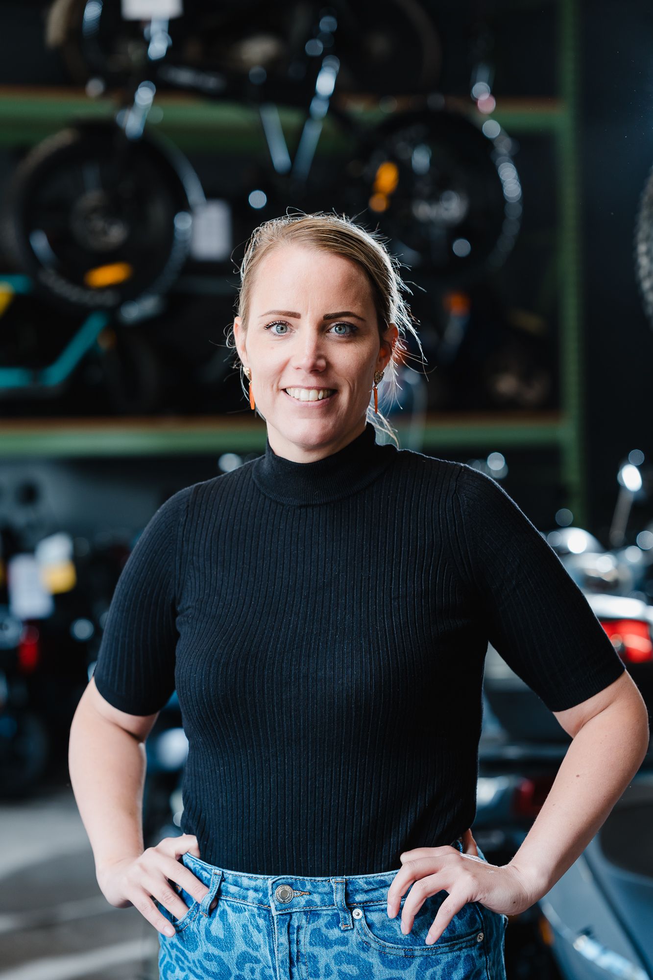 Portret van een vrouw in een werkplaats met fietsen op de achtergrond, handen in de zij.