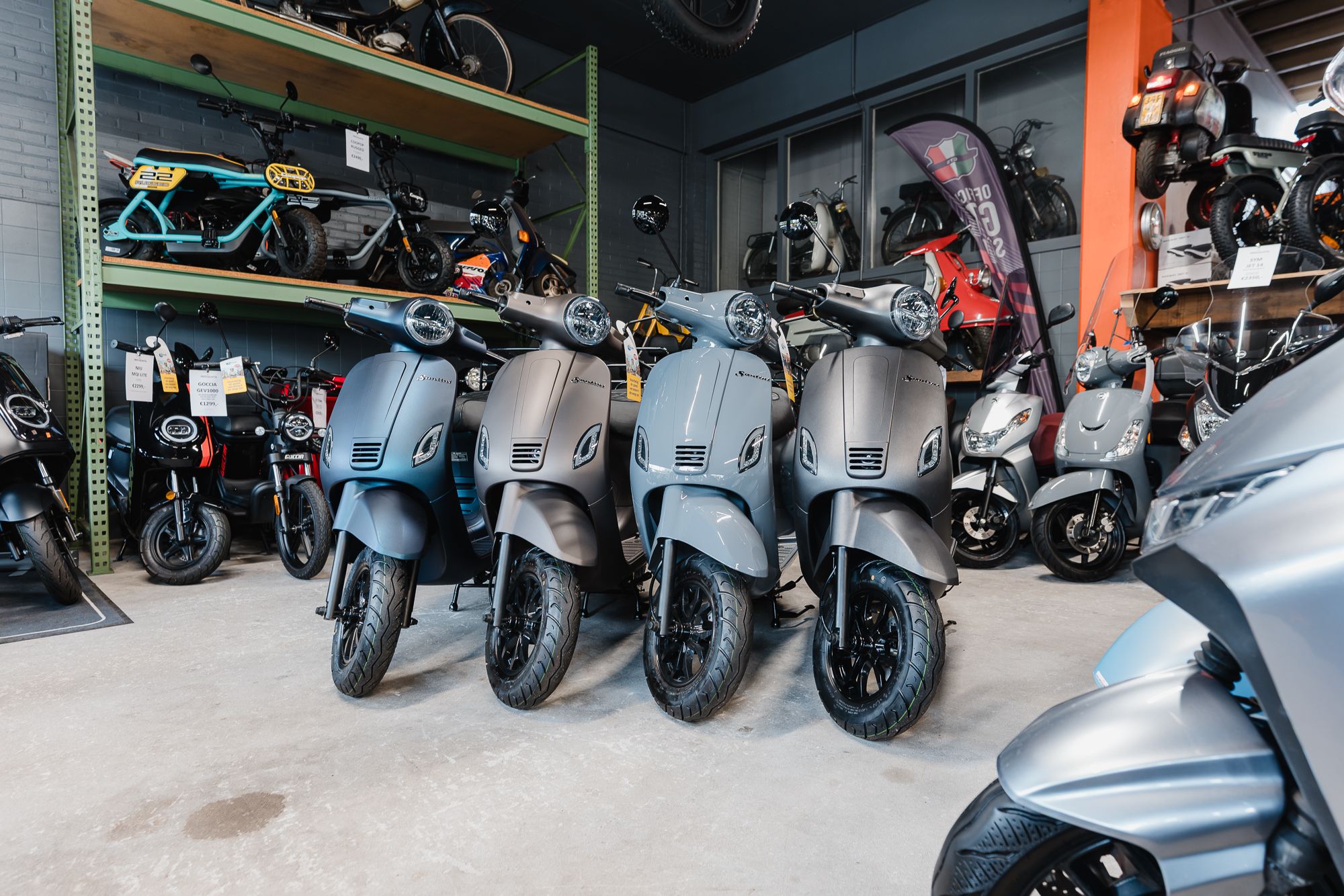 Vier grijze scooters op een rij in een showroom, omringd door andere brommers en accessoires.