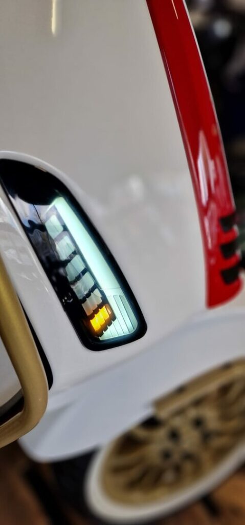 Detail van wit voertuig met verticale ledlamp, rode accentstrip en gouden spaakwiel.