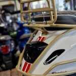 Achterkant van een crème scooter met goud bagagerek, rood-witte striping en blauw kenteken in showroom.