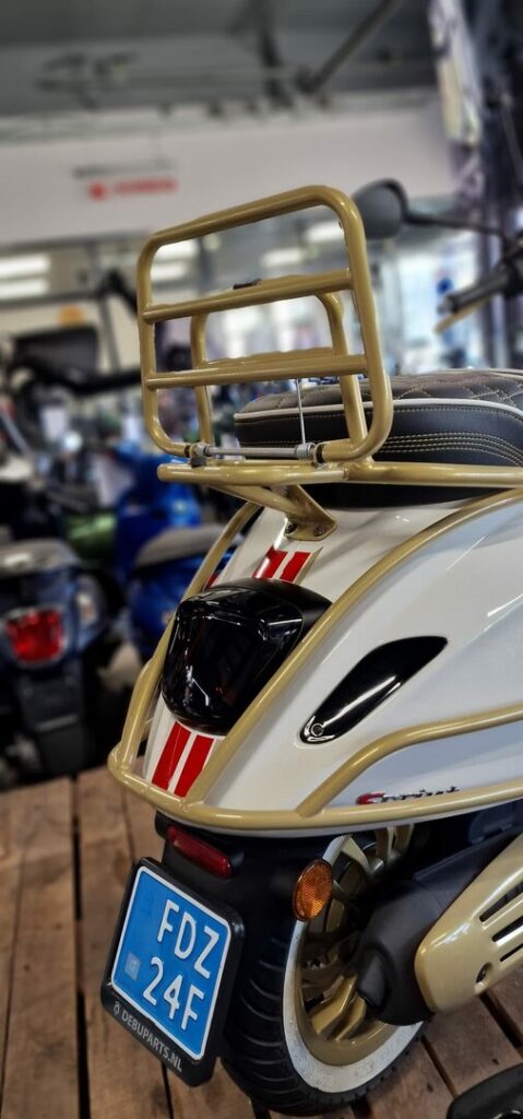 Achterkant van een crème scooter met goud bagagerek, rood-witte striping en blauw kenteken in showroom.