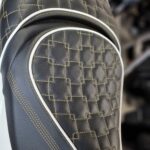 Close-up van een zwart motorzadel met ruitstiksel, goudkleurig garen en witte bies.