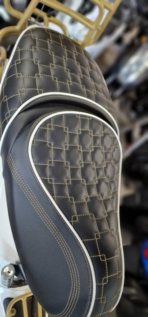 Close-up van een zwart motorzadel met ruitstiksel, goudkleurig garen en witte bies.