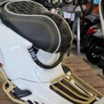 Close-up van een witte scooter met zwart doorgestikt zadel en gouden rekken in een werkplaats.