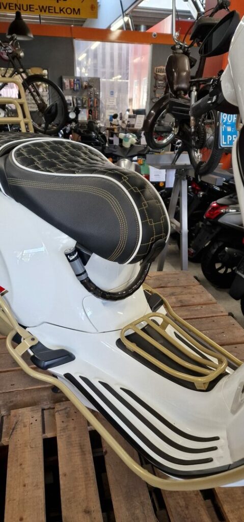 Close-up van een witte scooter met zwart doorgestikt zadel en gouden rekken in een werkplaats.