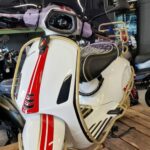 Witte Vespa met rode striping, hoog windscherm en goudkleurige valbeugels, gepresenteerd op houten pallets in een showroom.