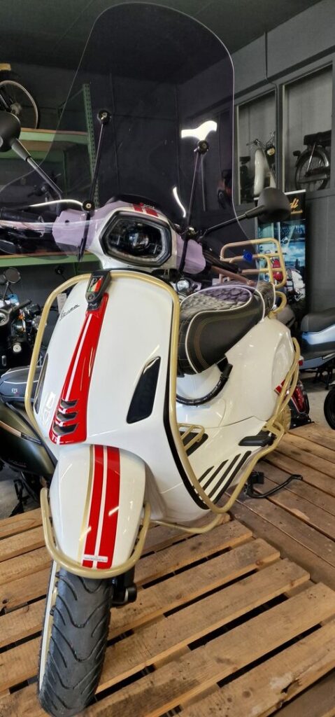 Witte Vespa met rode striping, hoog windscherm en goudkleurige valbeugels, gepresenteerd op houten pallets in een showroom.