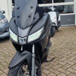 Zwarte motorscooter met hoog windscherm voor een garage, op klinkerstraat geparkeerd.
