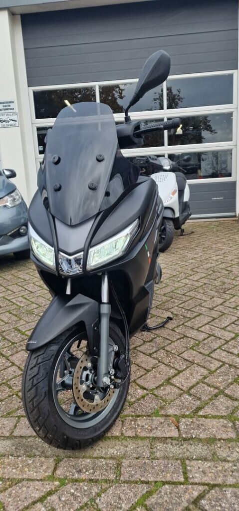 Zwarte motorscooter met hoog windscherm voor een garage, op klinkerstraat geparkeerd.