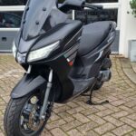 Zwarte motorscooter met hoog windscherm op de zijstandaard, geparkeerd op klinkers voor een garage