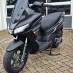 Mat zwarte motorscooter met hoge ruit geparkeerd op klinkers voor een garagedeur.