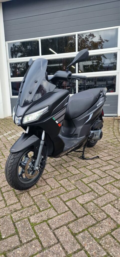 Mat zwarte motorscooter met hoge ruit geparkeerd op klinkers voor een garagedeur.