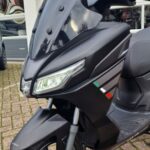 Close-up van een matzwarte scooter met groot windscherm, LED-koplamp en schijfrem bij een garage.