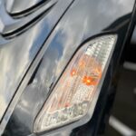 Close-up van brandend knipperlicht in koplamp van glanzend zwart voertuig met wolkenreflectie.