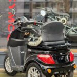 Zwarte driewielige scootmobiel geparkeerd voor een winkelraam met KYMCO-logo.