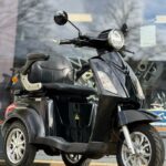 Zwarte driewielige elektrische scooter met rugleuning, geparkeerd voor een winkelraam op de stoep.