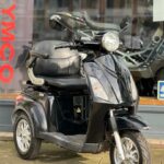 Zwarte driewielige scootmobiel met rugleuning en grote koplamp, geparkeerd voor een showroom.