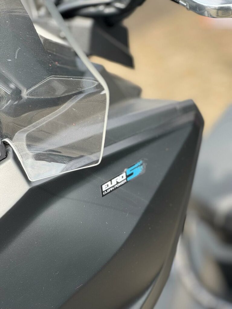 Close-up van een motorscooter met windscherm en Euro 5 Clean Power-sticker op matte kuip.