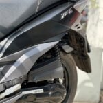 Close-up van de zijkant van een zwarte scooter met zadel, motorafdekking, achterwiel en vering.