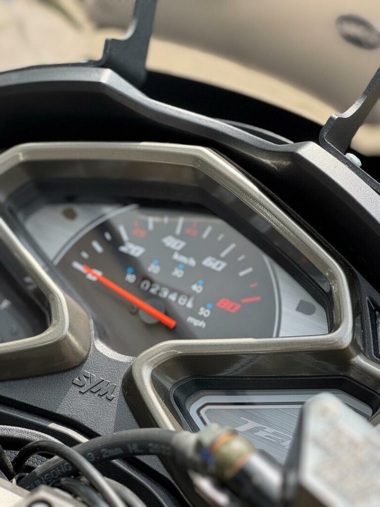 Close-up van het dashboard van een SYM-scooter met analoge snelheidsmeter en kilometerteller.