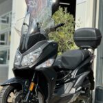 Zwarte maxiscooter met hoge ruit en topkoffer, geparkeerd buiten bij een werkplaats.