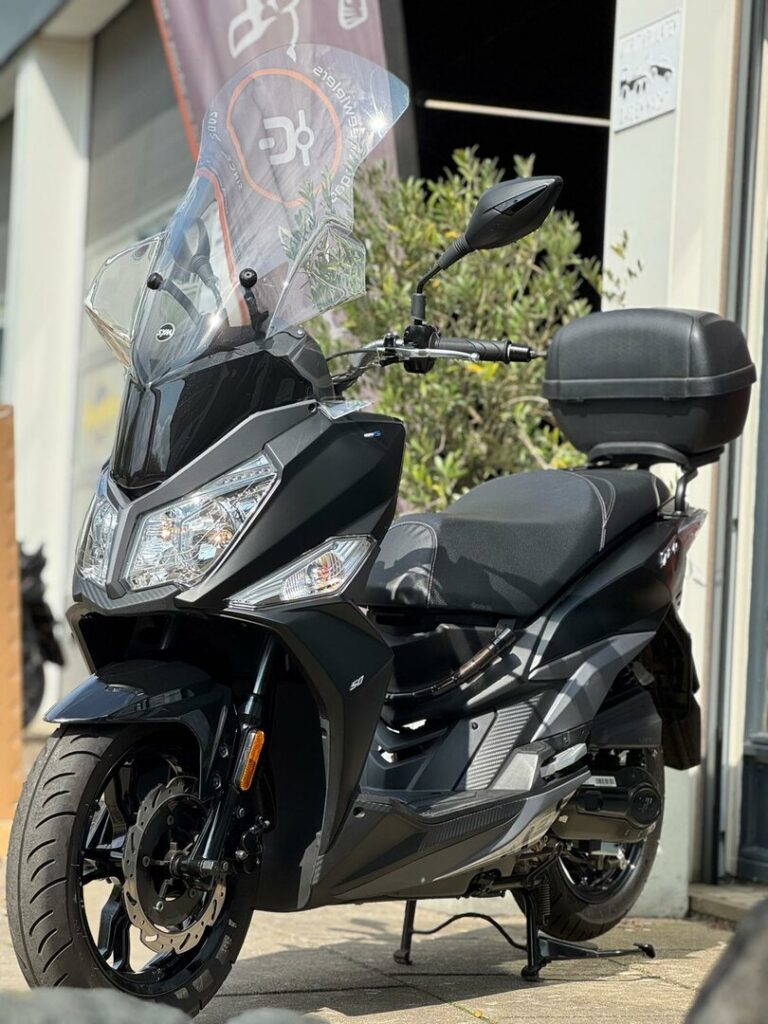 Zwarte motorscooter met hoog windscherm en topkoffer, geparkeerd voor een werkplaats in de zon.