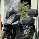 Zwarte moderne scooter met hoog windscherm en topkoffer, geparkeerd voor een werkplaats.