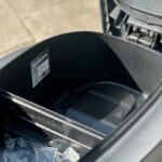 Geopende opbergruimte onder het zadel van een scooter, met sticker en plastic zakje.