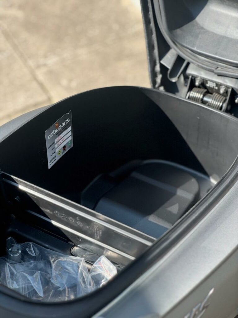 Geopende opbergruimte onder het zadel van een scooter, met sticker en plastic zakje.