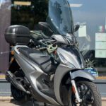 Zilveren motorscooter met hoog windscherm en topkoffer, geparkeerd voor een showroom.