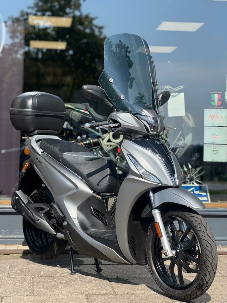 Zilveren motorscooter met hoog windscherm en topkoffer, geparkeerd voor een showroom.