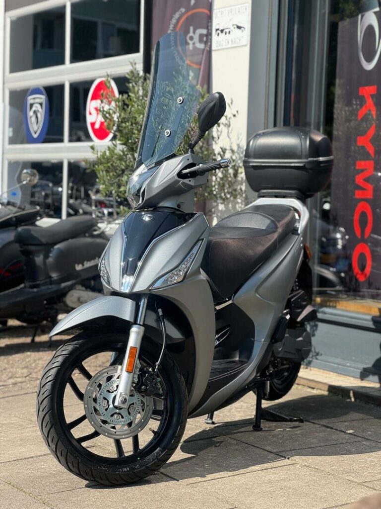 Zilverkleurige scooter met hoog windscherm en topkoffer, geparkeerd voor een scootershop.