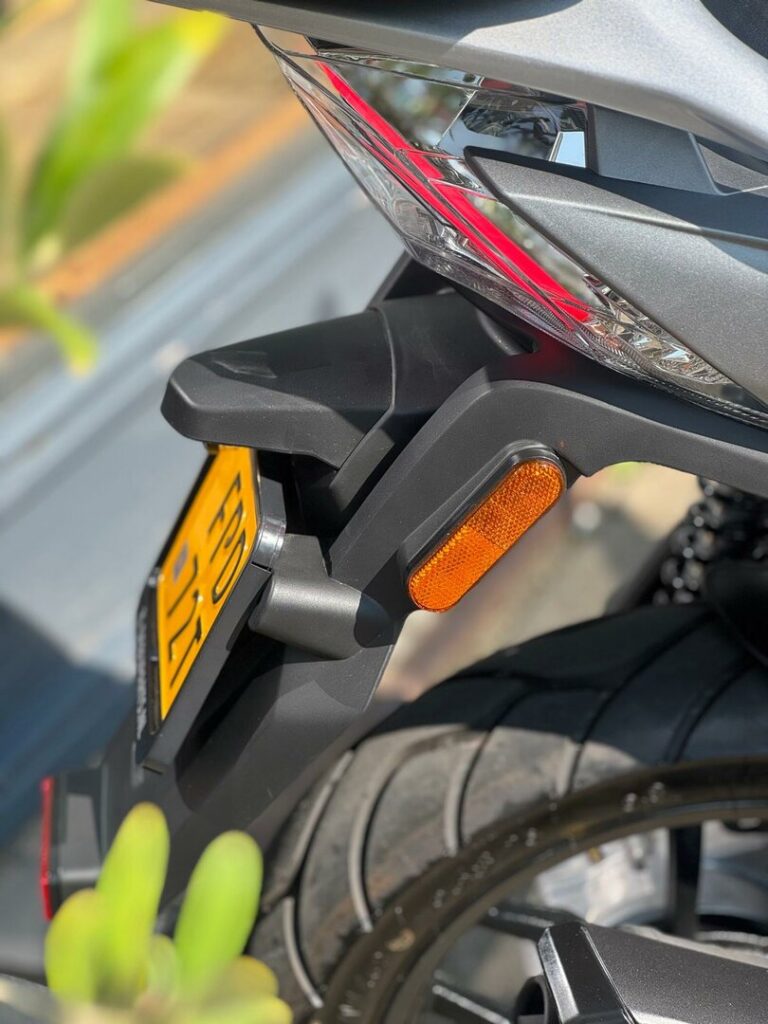 Close-up van het achterwiel van een motorfiets met geel kenteken, knipperlicht en achterlicht.