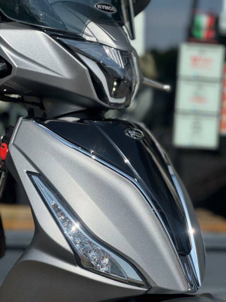 Close-up van de voorzijde van een zilveren KYMCO-scooter met scherpe LED-koplampen.