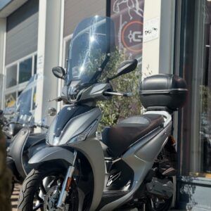 Zilveren motorscooter met hoog windscherm en topkoffer voor een showroom.