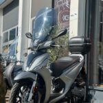 Zilveren stadsscooter met hoog windscherm en topkoffer voor een showroom op zonnige dag.