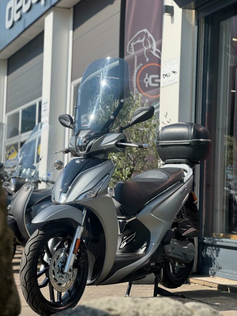 Zilveren stadsscooter met hoog windscherm en topkoffer voor een showroom op zonnige dag.