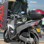 Grijze Kymco-scooter met hoog windscherm en topkoffer, geparkeerd voor een showroom.