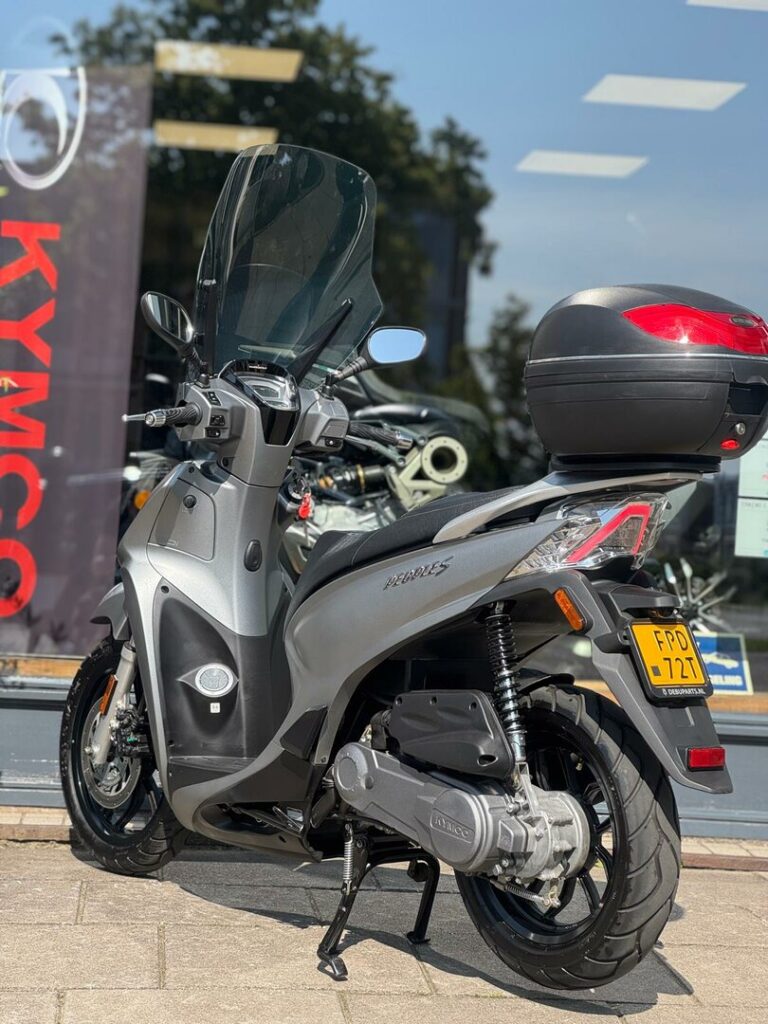Grijze Kymco-scooter met hoog windscherm en topkoffer, geparkeerd voor een showroom.