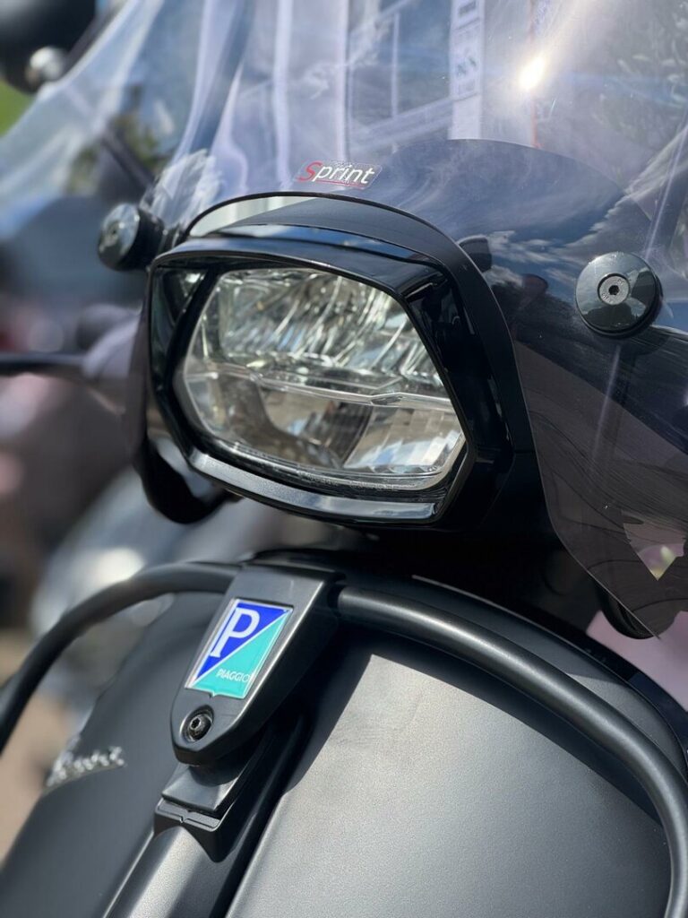 Close-up van de koplamp en het windscherm van een Vespa Sprint scooter met Piaggio-logo.