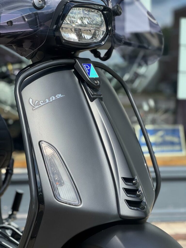 Close-up van de voorkant van een matte grijze Vespa-scooter met koplamp en ventilatieroosters.