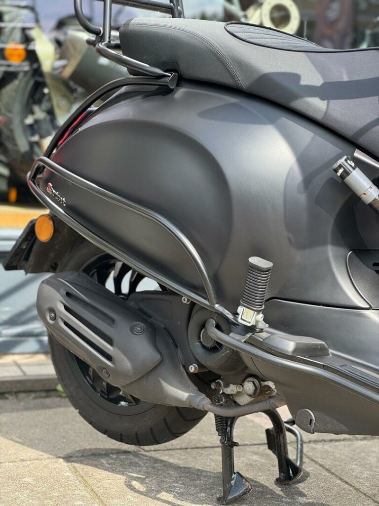 Close-up van de achterzijde van een matzwarte scooter met uitlaat, voetsteun en middenstandaard.