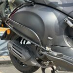 Close-up van een matzwarte scooter met uitlaat, achterwiel en middenbok