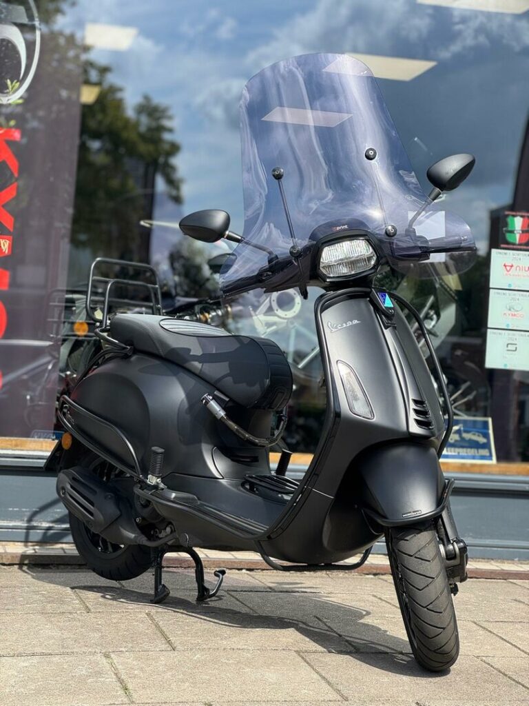 Matte zwarte Vespa-scooter met hoog windscherm geparkeerd voor een winkel
