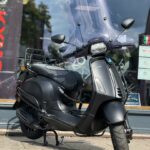 Mat zwarte Vespa-scooter met hoog windscherm, op standaard geparkeerd voor een winkelraam.