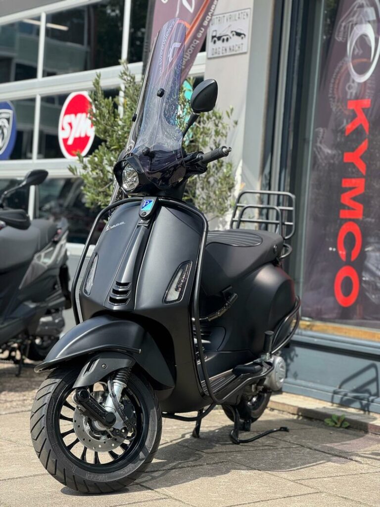 Matzwarte scooter met hoog windscherm, geparkeerd voor een scooterwinkel met banners.