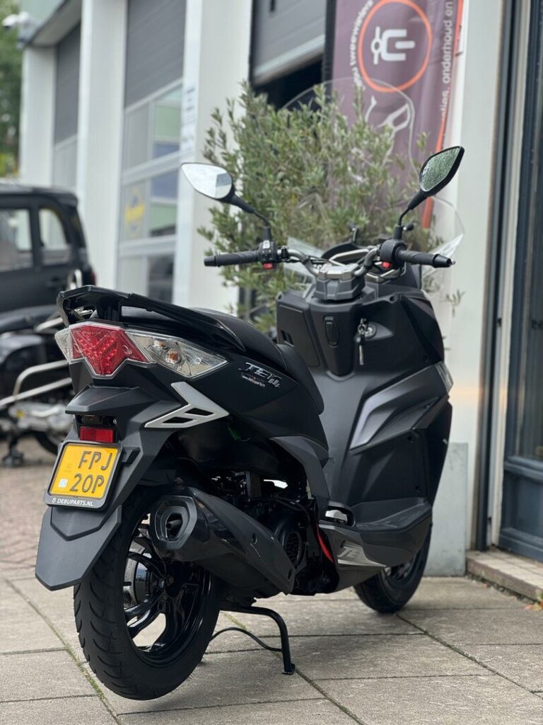 Zwarte scooter op middenbok met geel NL-kenteken voor een werkplaats.