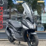 Zwarte motorscooter met hoog windscherm en LED-koplampen, geparkeerd voor showroom.
