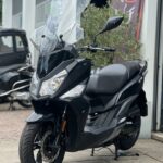 Zwarte motorscooter met hoge ruit op middenbok, geparkeerd voor een werkplaats.