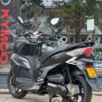Grijze Kymco-scooter met hoog windscherm, op standaard geparkeerd voor een showroom.